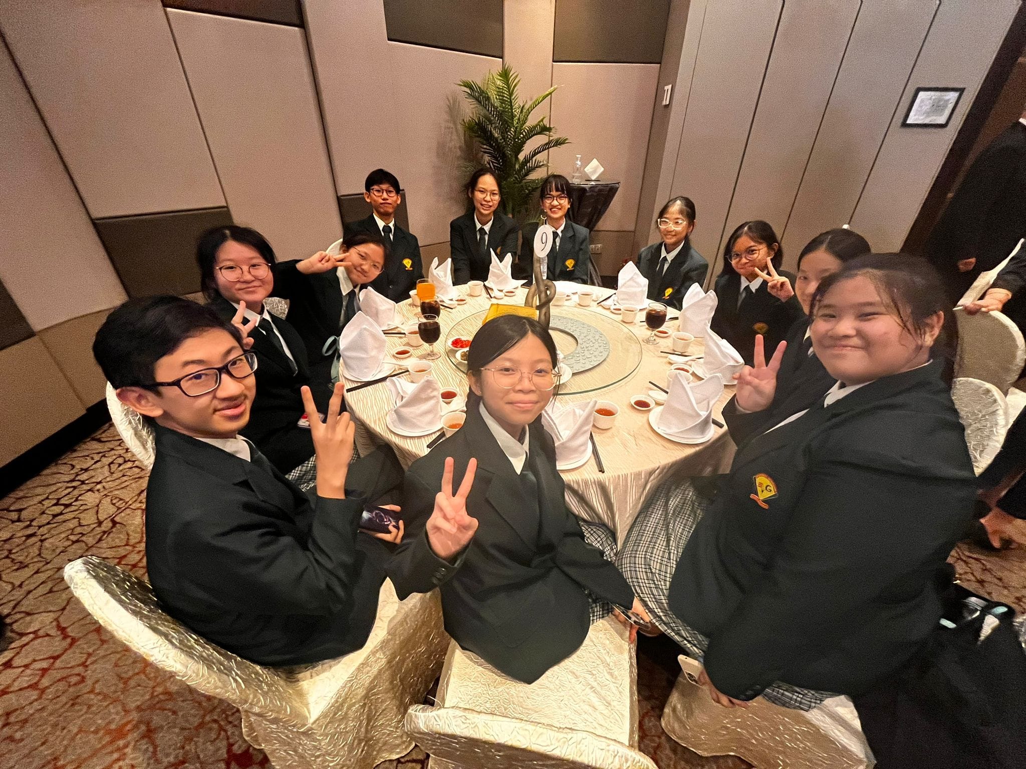 A group of students in dark uniforms sit around a round banquet table, smiling and making peace signs.